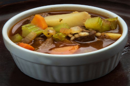 A vegetable soup garnished with pieces of carrots and pieces of green onion, and selery, served in a white bowl.